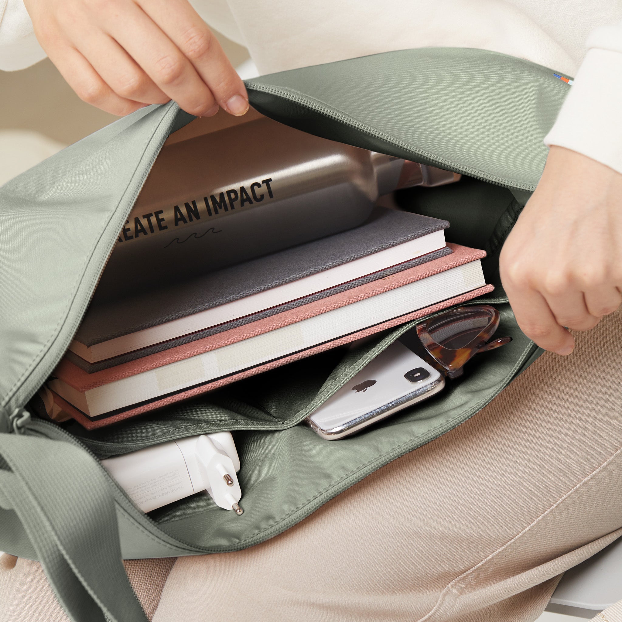 Eine Person öffnet die MOON BAG LARGE Tasche in bass (Salbeigrün) mit Büchern und persönlichen Gegenständen.