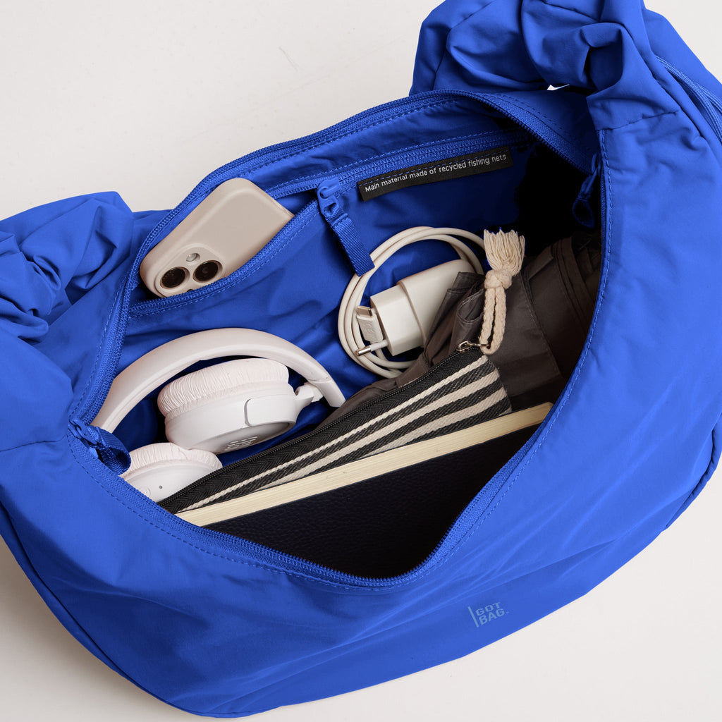 MOON BAG RUFFLE Tasche in cobalt (leuchtendes Kobaltblau), geöffnet mit Inhalt wie Smartphone und Kopfhörer.
