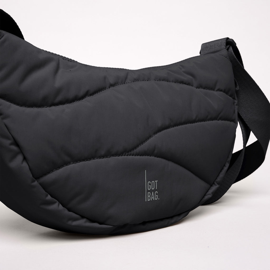 WAVY PUFFER MOON BAG Tasche in black (Schwarz) mit wellenförmiger Steppung auf weißem Hintergrund.