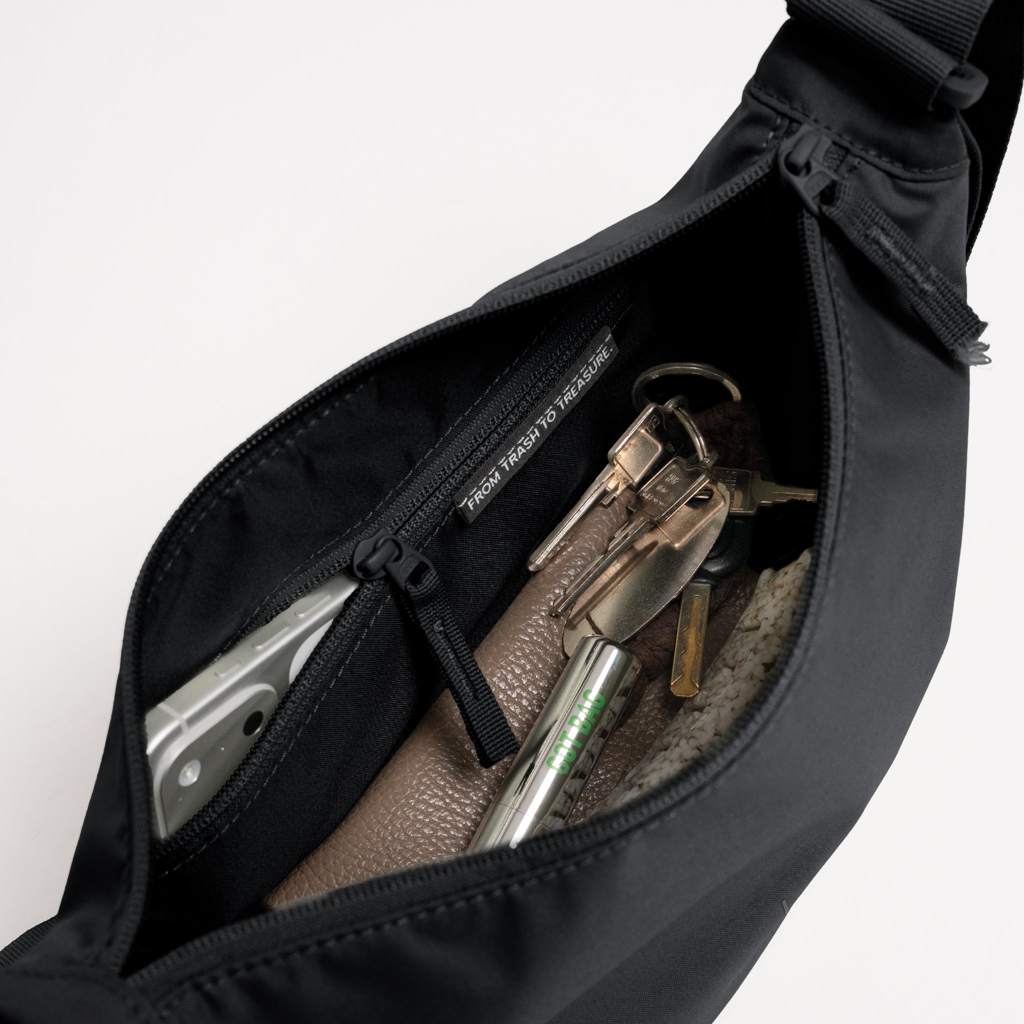 MOON BAG SMALL Tasche in black (Schwarz) mit offenem Innenraum, der Smartphone, Schlüssel und eine FROM TRASH TO TREASURE Aufschrift zeigt.